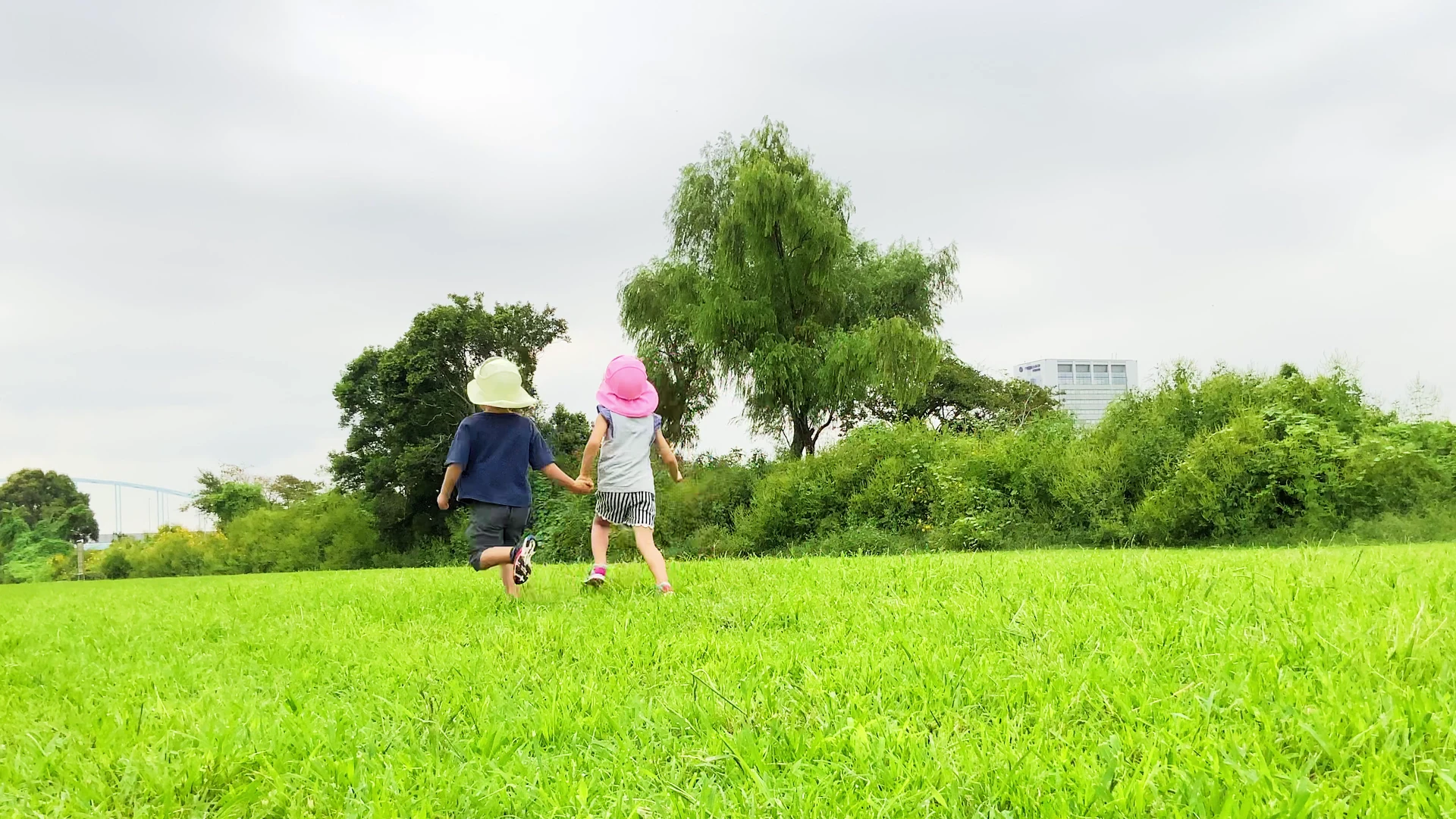 クリーム色とピンクのまもる君をかぶり、手をつないで芝生を走る2人の子ども